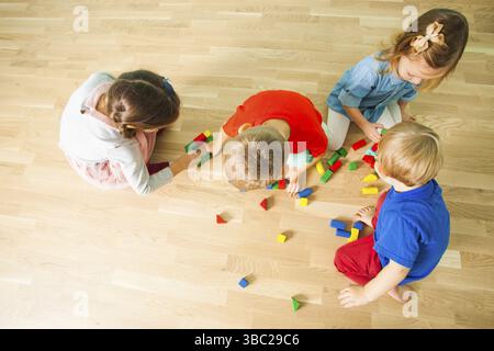 Four kids playing on a wooden floor with colorful wooden bricks. Top view photo of children in kindergarten building toy towers. Free game concept Stock Photo