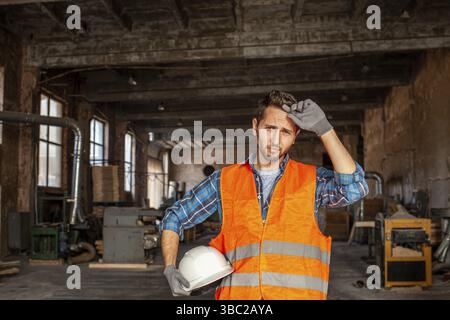 Portrait of young handsome engineer feeling tired after work, standing indoors his working place at the shop of woodworking plant. Male worker with ha Stock Photo