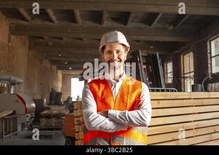 Young smiling and happy engineer in orange vest and hard hat. Wood worker indoors at factory Stock Photo