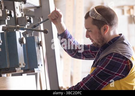 Closeup back view young professional woodworker in yellow vest, holding hydraulic press lever operating it to glue together pieces of wood. Woodworkin Stock Photo