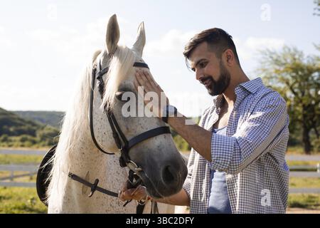 The young smiling man is standing next to a white horse and stroking it the head. The bearded man with horse in a ranch Stock Photo