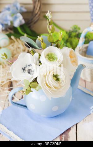 Close view of spring bouquet of large white ranunkulus flowers in blue teapot with white dots. Teapot with flowers on blue napkin on wooden surface Stock Photo