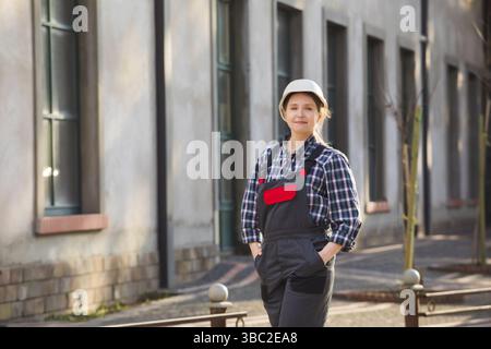 Portrait of happy, self-confident female worker in white hard hat and working uniform, posing with hands folded on chest. Young professional is proud Stock Photo
