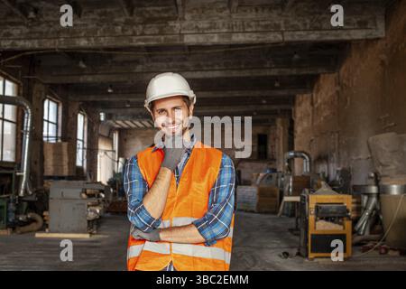 Portrait of happy young foreman with hard hat and protective gloves, standing indoors old factory, holding his chin, smiling, looking at the camera Stock Photo
