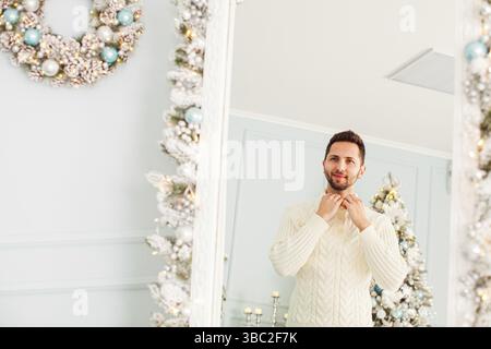 Young handsome man in a white knitted sweater looking at himself in large mirror at home decorated for Christmas. Reflection in the mirror, winter hol Stock Photo