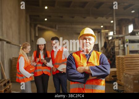 Team of young business people in orange protective vests and hard hats, standing in group on the background, mature foreman holding hands at chest, st Stock Photo