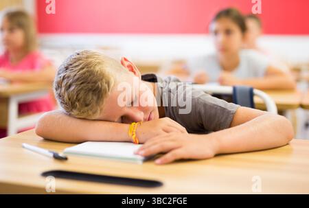 Bored tween boy sleeping at desk in classroom during lesson Stock Photo