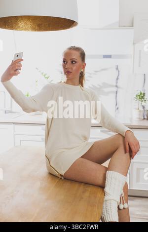 Profile side view of attractive and vogue young lady, in knitted white dress and kintted stockings with ponytail hairdo, taking selfie, sitting or lar Stock Photo