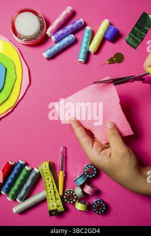 Child hands holding scissors and cutting out the felt. Colorful spools and bobbins, needles placed on the pink surface. Moment of kid's creativity Stock Photo