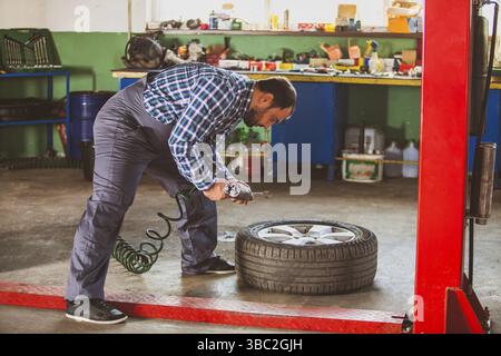 Side view of man, professional auto mechanic, repairing car wheel on a floor at car service workshop. Table with variety of tools on a background Stock Photo