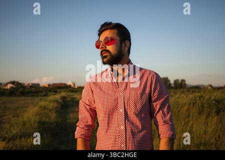 Happy young man posing outdoor on summer field background. Handsome bearded man in pink sunglasses enjoying summer sun outdoors in countryside Stock Photo