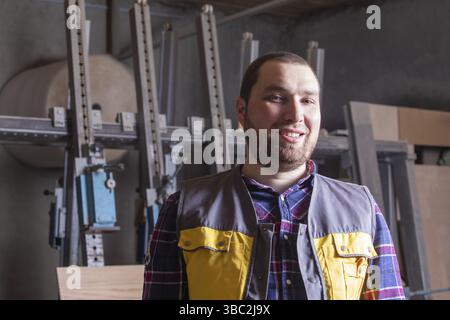 Portrait of young smiling satisfied bearded carpenter feeling very proud to be member of team. Woodworking factory worker at his working place. Loving Stock Photo