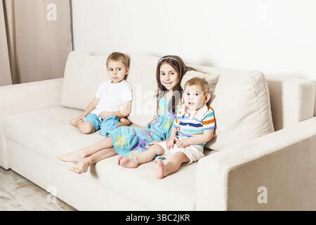 Three children sitting on a light sofa. Sister and her two younger brothers in the living room Stock Photo