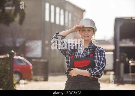 Portrait of happy, self-confident female worker in white hard hat and working uniform, posing with hands folded on chest. Young professional is proud Stock Photo