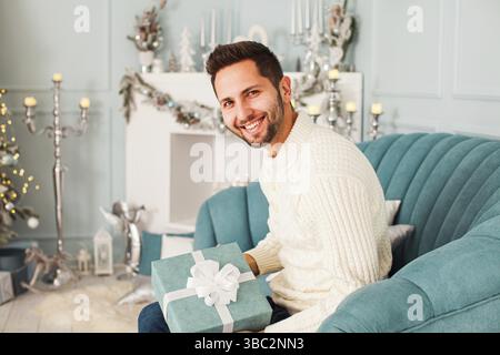 Young attractive smiling man in white sweater sitting on a blue couch and looking at the camera, holding blue gift box with a white ribbon. Christmas Stock Photo