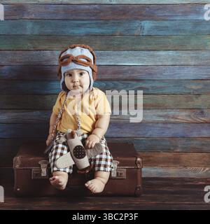 Boy in a knitted pilots cap plays with toy airplane, sitting on an old-fashioned suitcase Stock Photo