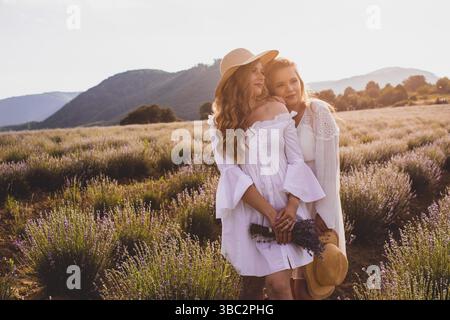 Summer sunset over two women friends in white dresses, standing close to each other in lavender field, looking away in back lit. Spending time togethe Stock Photo