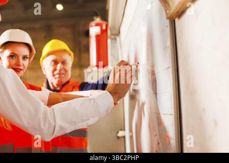 The experienced engineer make practices for beginners at the factory. The engineers are standing next by flip chat Stock Photo