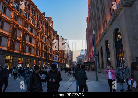 Shanghai, China - 1 April 2025: Tourists stroll along Nanjing Road at dusk with a glowing view of historic Bund buildings lit in warm golden light acr Stock Photo