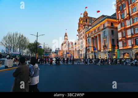 Shanghai, China - 1 April 2025: Tourists stroll along Nanjing Road at dusk with a glowing view of historic Bund buildings lit in warm golden light acr Stock Photo