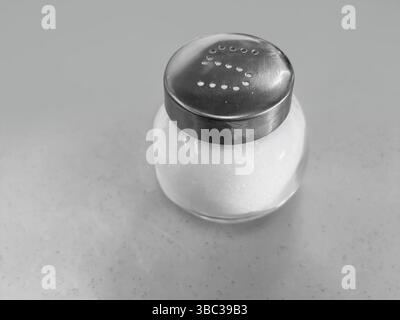 Minimalistic black and white image featuring a salt shaker with a metallic top and transparent body, placed on a smooth flat surface. Ideal for themes Stock Photo