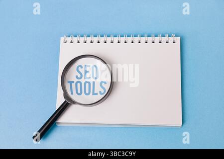 A magnifying glass with the words "SEO TOOLS" enlarged underneath rests on a blank notepad against a light blue background. Stock Photo