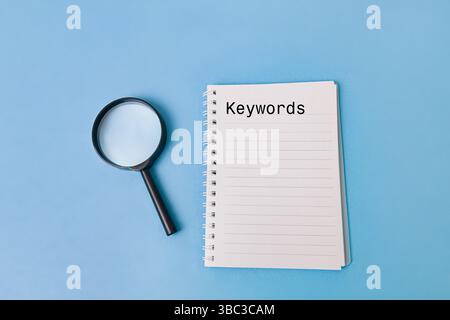 A magnifying glass sits beside a notepad with the word "Keywords" prominently displayed. This highlights the importance of keyword research in various Stock Photo