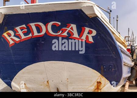 The stern of a fishing boat with large lettering viewed from a low-angle is situated near a sea wall.  A hazy blue sky is above. Stock Photo