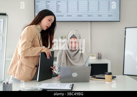 Two young Asian women, one wearing a hijab, collaborate on a laptop in a modern office. Discussing business strategy, financial planning, and teamwork Stock Photo