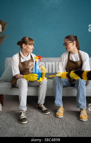 Two Female Janitors Sitting on Sofa Resting and Talking After Cleaning. communicating and relaxing after completing housework chores Stock Photo