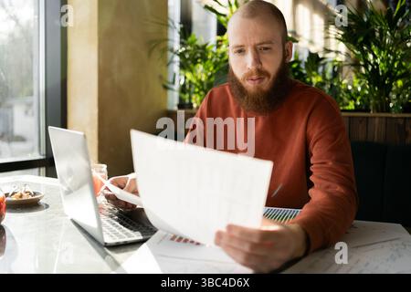 entrepreneurship. Bearded handsome man reading document sitting with laptop in cafe near window. Modern business startup and planning Stock Photo