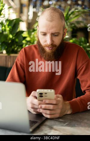 Millennial man using cellphone for chatting while sitting near laptop computer in office Stock Photo