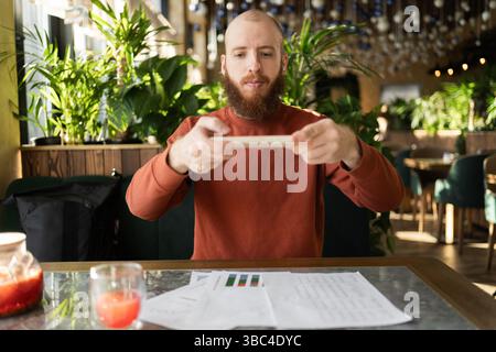 Concept of modern gadgets and business. Bearded businessman wearing sweater using phone, taking photo of documents and sending to client, sitting at Stock Photo