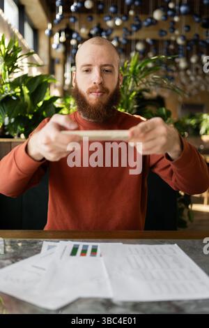 Modern gadgets and business concept. Young bearded businessman using phone, taking photo of documents sitting at desk in modern cafe Stock Photo