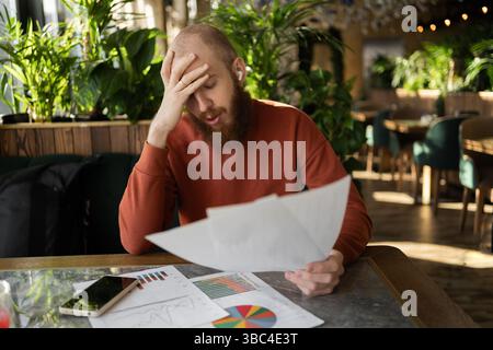 Young bearded accountant manager working remotely in cafe reading documents and holding head with hands, concept of problems and bankruptcy Stock Photo