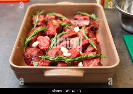 Uncooked beef pieces in a ceramic tray seasoned with fresh rosemary sprigs and garlic cloves, ready for oven roasting. Stock Photo