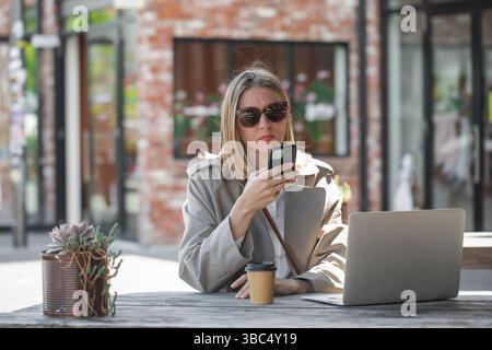 Young woman working using phone and laptop sitting on cafe veranda. Remote work, businesswoman Stock Photo