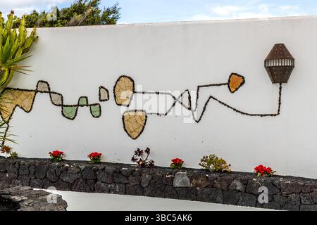 An abstract mosaic artwork made of dark tile fragments stretches across a white wall at the Fundación César Manrique in Tahíche, Lanzarote, with a hexagonal lantern at the right edge of the frame. Red flowers grow along the natural stone wall in front of it. César Manrique returned from New York in 1966 and built his home here, where he lived from 1968 to 1988 – longer than anywhere else in his life. Since 1992 it has been open to the public as a foundation. Calle Amando Fuentes, Teguise, Canary Islands, Spain. Stock Photo