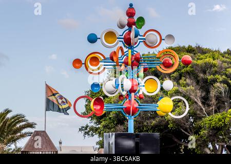 A colourful kinetic wind sculpture with spheres and rings in red, orange, yellow, blue and white turns in front of trees near the Fundación César Manrique in Tahíche, Lanzarote - possibly one of César Manrique's "Juguetes del Viento" (wind toys), several of which are installed across the island. Calle Amando Fuentes, Teguise, Canary Islands, Spain. Stock Photo