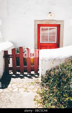 Bright red wooden door framed in stone, with a matching rustic gate leading through curved whitewashed walls, bathed in strong sunlight. Foreground pl Stock Photo