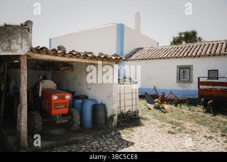 Traditional farmyard scene with a small red tractor parked under a tiled shed, surrounded by rustic tools, barrels, and whitewashed buildings with blu Stock Photo