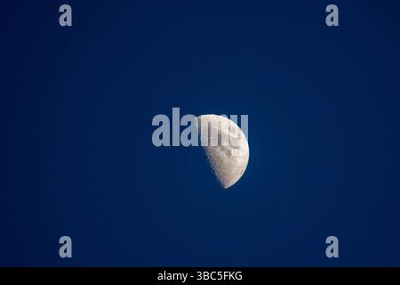 A half moon glows brightly against a vast, deep blue sky, capturing the serene beauty of space. Stock Photo