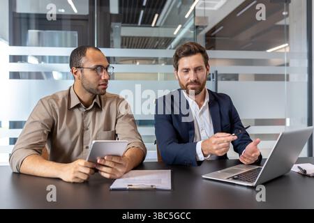 Two business colleagues collaborate, using a laptop and tablet to review documents and discuss ideas at their office table. Stock Photo