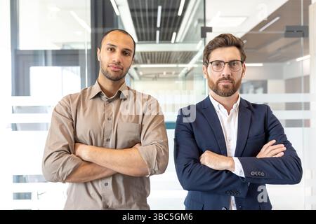 Two business professionals stand confidently, arms crossed, in a modern office setting. They look directly at the camera. Stock Photo