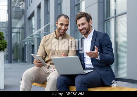 Two business partners sit outdoors, collaborating on a laptop and tablet, discussing work-related data. Stock Photo