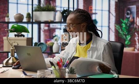 Teleworker using laptop in home office to crosscheck paper documents with online company files. Remote worker typing on notebook, inputting paperwork documentation data, camera B Stock Photo