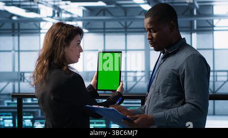 Computer scientists in data center use mockup tablet, inspecting server rigs, updating devices. Teamworking IT colleagues collaborating to ensure stable operations, using chroma key device, camera A Stock Photo