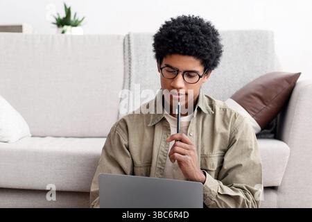 Focused black teen guy sitting on floor with study materials and laptop, at home Stock Photo
