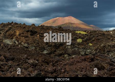 Volcanic landscape near El Golfo on Lanzarote. Dark lava rock formations cover the slope, on which small plants have colonised. A reddish volcanic cone can be seen in the background, overlaid by dark clouds. The scenery shows the power of nature and the adaptability of life in volcanic areas. Avenida Marítima, El Golfo, Canarias, Spain Stock Photo