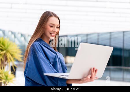 Smiling businesswoman in blue shirt using laptop at workplace. Beautiful woman in smart casual working on netbook, focused and engaged in professional environment with modern office setting Stock Photo